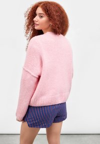 Übergroßer rosa Strickpullover mit strukturiertem Finish und geripptem Saum, kombiniert mit navy-blauen gestreiften Shorts. Der Pullover hat fallende Schultern.