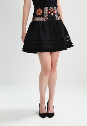 Kobieta w czarnej plisowanej spódnicy z kolorową haftowaną talią i czarnych szpilkach, stojąca na gładkim jasnym tle.