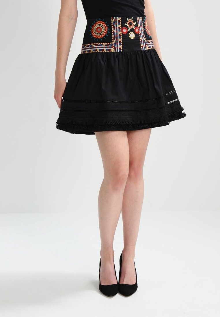 Femme portant une jupe plissée noire avec une ceinture brodée colorée et des escarpins noirs, debout devant un fond clair uni.