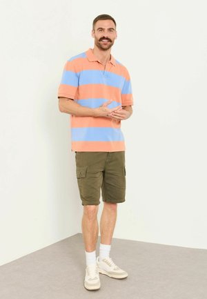 Mann steht drinnen und trägt ein kurzärmeliges, orange-blau gestreiftes Polo, olivfarbene Cargoshorts, weiße Socken und weiße Turnschuhe.