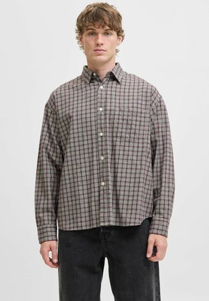 Young man wearing a loose, long-sleeve checked shirt in red and white paired with black jeans, standing against a plain white background.