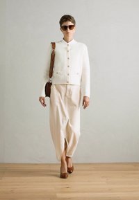 Veste blanche texturée avec des boutons dorés, associée à un pantalon large beige clair et des talons marron. Les accessoires incluent de grandes lunettes de soleil et un sac marron.