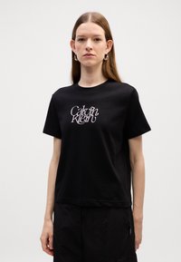 T-shirt en coton noir avec col rond et manches courtes, présentant un logo "Calvin Klein" en cursive blanc sur le devant.