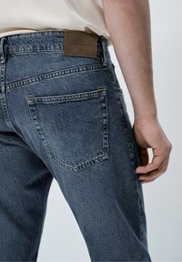 Mörkblå denimjeans har en bakficka och en brun läderetikett vid midjan. Tyget har en bleknad textur.
