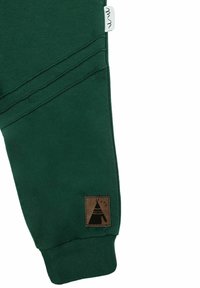 Ciemnozielone spodnie dresowe o fakturowanym wykończeniu, z brązową tkaną metką przedstawiającą czarne tipi na dolnej lewej stronie.