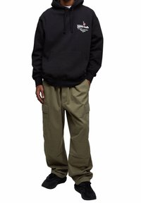 Mężczyzna w czarnej bluzie z kapturem z logo i napisem na piersi, oliwkowo-zielonych spodniach typu cargo i czarnych trampkach, stoi z jedną ręką w kieszeni.