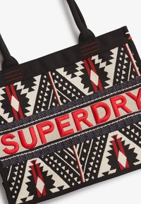 Borsa tote nera con motivi geometrici in rosso e bianco. Presenta un logo "SUPERDRY" rosso ben in evidenza, accentuato da dettagli blu navy.