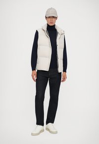 Hellgraue gepolsterte Weste über einem dunklen Rollkragenpullover, kombiniert mit dunklen Jeans und weißen Sneakers. Eine hellgraue Kappe rundet das Outfit ab.