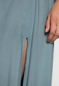 Lichtblauwe jurk van zachte stof, met een split aan de zijkant en een gladde textuur. Inclusief een close-up van een hand die tegen de stof rust.