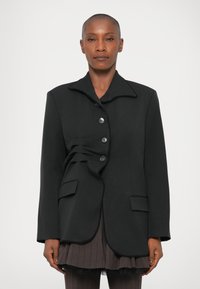 Blazer negro a medida con dobladillo curvado, cuello mao y cierre de cinco botones. Presenta un detalle plisado único y dos bolsillos frontales.