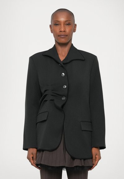 Schwarzer maßgeschneiderter Blazer mit geschwungenem Saum, Mandarin-Kragen und fünf Knöpfen. Verfügt über einzigartige Faltendetails und zwei Fronttaschen.