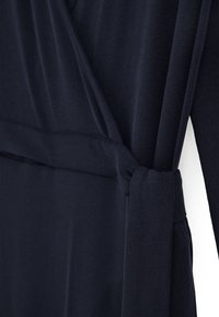 Détail d'une robe portefeuille en tissu bleu marine foncé avec un nœud noué à la taille, montrant une texture lisse et une manche longue.