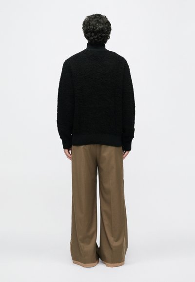 Schwarzer strukturierten Pullover mit hohem Kragen, kombiniert mit weit geschnittenen beige-braunen Hosen, von hinten vor einem schlichten weißen Hintergrund betrachtet.
