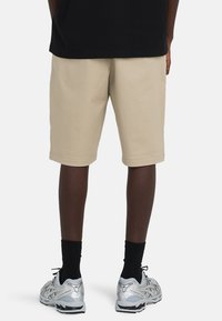 Vista trasera de una persona con pantalones cortos beige hasta la rodilla, calcetines negros, zapatillas de correr plateadas y camiseta negra sobre un fondo liso.