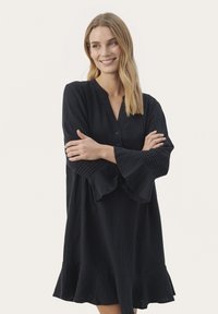 Rochie neagră, fluidă, cu decolteu în V, închidere cu nasturi și mâneci evazate. Prezintă un tiv volănat și detalii texturate pe mâneci.