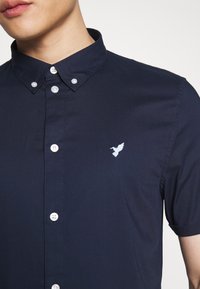 Uomo che indossa una camicia button-down blu navy con maniche corte e un piccolo logo a forma di uccello bianco ricamato sul lato sinistro del petto.