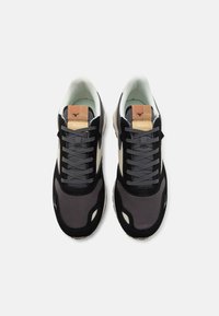 Black and gray trainers with mesh and suede materials, featuring a tan tongue label, lace-up design, and contrasting white accents.