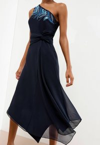 Nais wearing a navy blue one-shoulder dress with blue sequin feather detail and knotted waist, flowing asymmetrical hem.