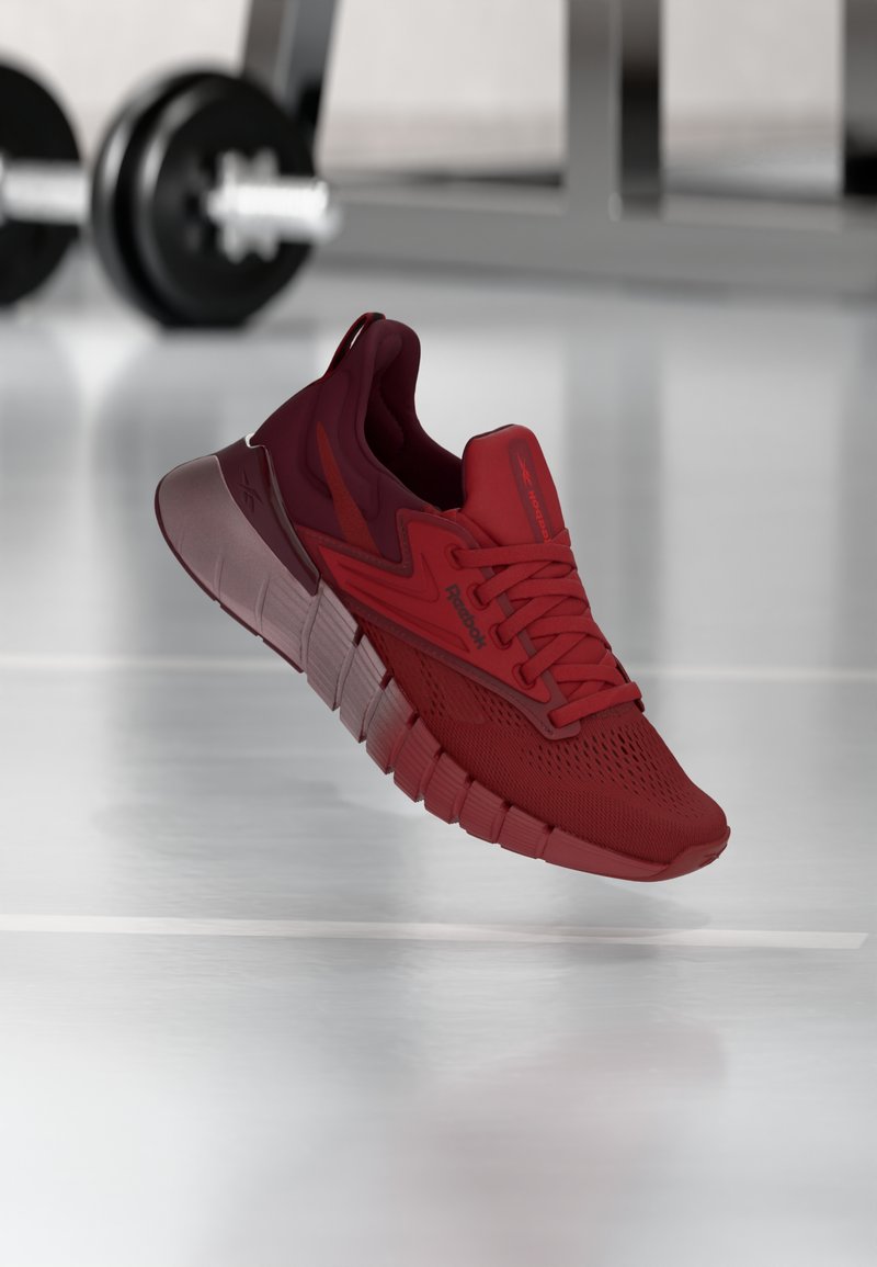 Κόκκινο αθλητικό παπούτσι Reebok κρεμασμένο πάνω από το δάπεδο του γυμναστηρίου, με θολά αλτήρα και πάγκο προπόνησης στο φόντο.