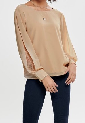 Femme portant un chemisier beige avec des panneaux en dentelle transparente sur les manches longues et un pantalon foncé ajusté, debout, mains près des hanches.