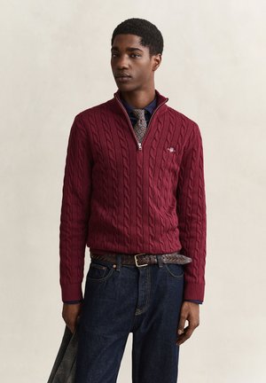 Junger Mann trägt einen burgunderfarbenen, kabelgestrickten Quarter-Zip-Pullover, gemusterten Krawatte, dunkelblaue Jeans und hält eine graue karierte Jacke in einer Hand.