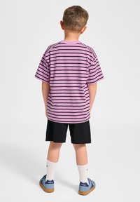 Lila und schwarz gestreiftes T-Shirt mit kurzen Ärmeln, kombiniert mit schwarzen Shorts. Getragen mit weißen Socken und blauen Schuhen, von hinten gesehen.