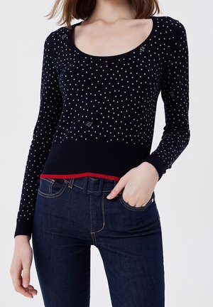 Vrouw draagt een marineblauwe trui met polkadots en rode biezen en donkerblauwe hooggetailleerde spijkerbroek, hand in zak tegen een witte achtergrond.