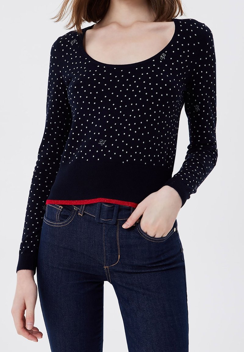 Femme portant un pull bleu marine à pois avec bordure rouge et un jean taille haute bleu foncé, main dans la poche sur fond blanc.