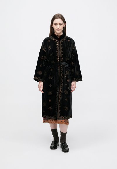 Kvinde stående iført en sort broderet fløjlsrobe bundet i taljen over en brun kjole med mørke sokker og sorte loafers på hvid baggrund.
