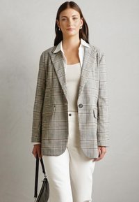 Blazer grigio a quadri con vestibilità su misura, due tasche frontali e chiusura con un bottone, abbinato a un top bianco e pantaloni crema.