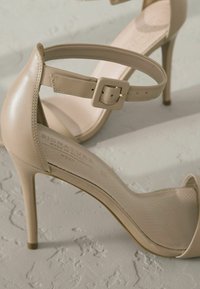 Sandales à talons hauts beige avec une tige en cuir lisse, une sangle de cheville avec une boucle carrée et une semelle intérieure texturée arborant un logo de marque.