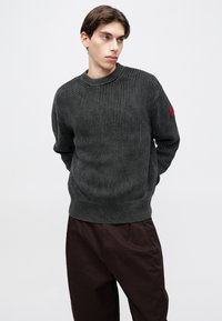 Suéter de punto gris oscuro con diseño acanalado, escote redondo y detalle rojo en la manga, combinado con pantalones marrones de corte holgado.