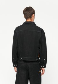 Chaqueta de mezclilla negra con un diseño clásico, que presenta un collar puntiagudo, cierre frontal con botones y bolsillos laterales. Textura suave con costuras sutiles.