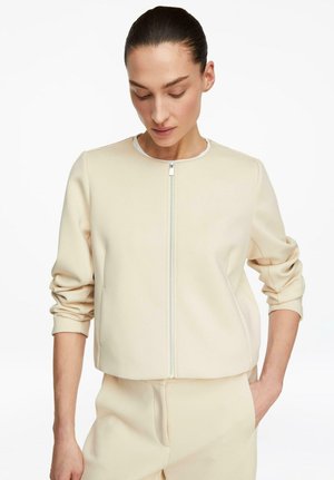 Woman with dark hair tied back wearing a cream zip-up jacket and matching pants, looking down with one hand in pocket.