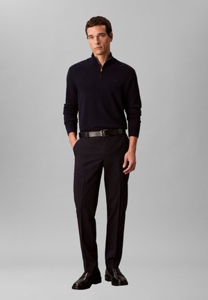 Mann stående med hendene i lommene iført svart kvartglidelås-sweater, svarte bukser, svart skinnbelte og svarte sko mot grå bakgrunn.