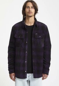 Chemise en flanelle à carreaux noirs et violets avec manches longues, boutons à l'avant, deux poches poitrine et un ourlet arrondi, portée par-dessus un t-shirt noir.