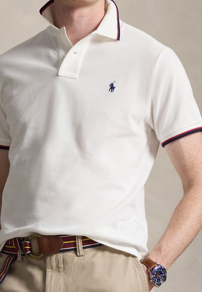 Férfi fehér galléros pólót visel tengerészkék és piros szegéllyel, bézs nadrágot, csíkos övet és barna karórát kék számlappal.