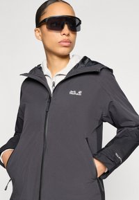 Veste imperméable noire avec capuche, fermeture éclair à l'avant et poches latérales. Portée sur un haut clair, accompagnée de grandes lunettes de soleil foncées.