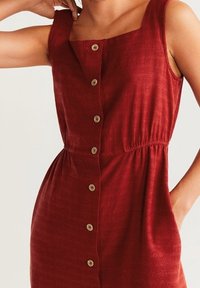 Robe en lin rouge avec décolleté carré, boutons sur le devant, taille élastique pour la silhouette et poches sur les côtés. Texture lisse, design décontracté.