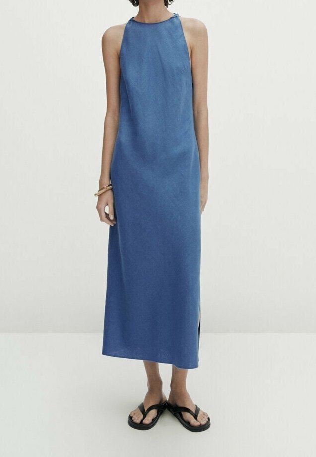 Femme portant une robe bleue sans manches, longue jusqu'aux chevilles, avec une fente latérale, des sandales noires à lanières et un bracelet au poignet gauche.