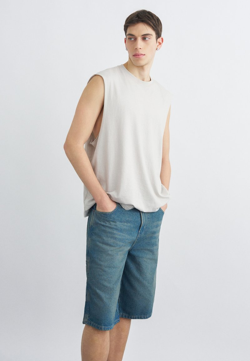 Jeune homme aux cheveux courts et foncés, portant un débardeur blanc et un short en jean bleu jusqu'aux genoux, debout avec les mains dans les poches.