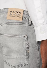Jeans in denim grigio con un'etichetta in pelle che riporta "SCOTCH & SODA AMSTERDAM." Presentano dettagli di cucitura a contrasto vicino alla tasca.