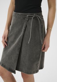 Harmaa denim-kietaisuhame, jossa on solmittava vyö vyötäröllä, yhden sivutaskun ja suora helma. Pehmeä rakenne ja minimisetomukset.