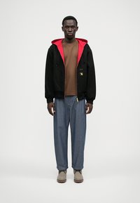 Črni pulover z zadrgo in rdečo podlogo, pod njim rjava srajca, ohlapne modre kavbojske hlače in siva obutev. Minimalistična zasnova z žepi.