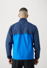 Giacca a vento con parte superiore blu navy e parte inferiore blu brillante. Tessuto liscio, colletto rialzato, polsini elasticizzati e vestibilità comoda.