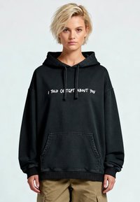 Felpa nera con cappuccio con scritta bianca, tasca anteriore e polsini a costine. Caratterizzata da un design ampio e una texture morbida, adatta per un abbigliamento casual.