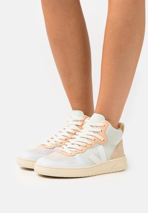 Jambes portant des baskets montantes en blanc, beige et tons pêche avec des lacets blancs et une semelle beige sur un fond uni.