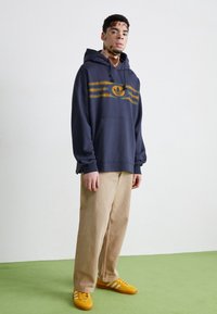 Marinblå hoodie med guldstripes och logotyp, i en kombination med beigefärgade vida byxor och orangea sneakers. Enkel design och avslappnad passform.