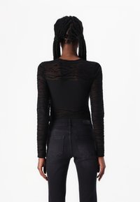 Langärmliges schwarzes Oberteil mit einem strukturierten, transparenten Rücken und einer soliden Vorderseite, kombiniert mit dunklen Jeans im klassischen fünfteiligen Design.