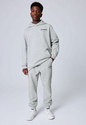 Ung mand står iført lysegrå AllSaints hoodie og matchende joggingbukser med hvide sneakers mod ensfarvet baggrund.
